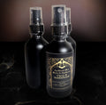 Three matte black glass spray bottles on a dark marble surface, the front bottle showing the gold Clove & Sandalwood label.