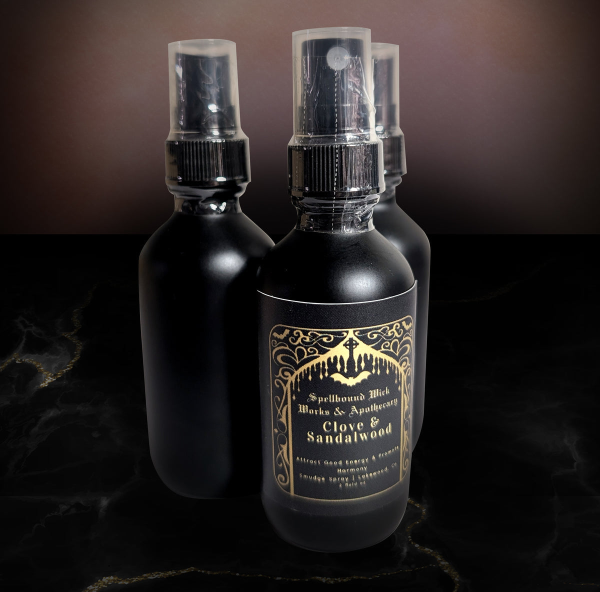 Three matte black glass spray bottles on a dark marble surface, the front bottle showing the gold Clove & Sandalwood label.