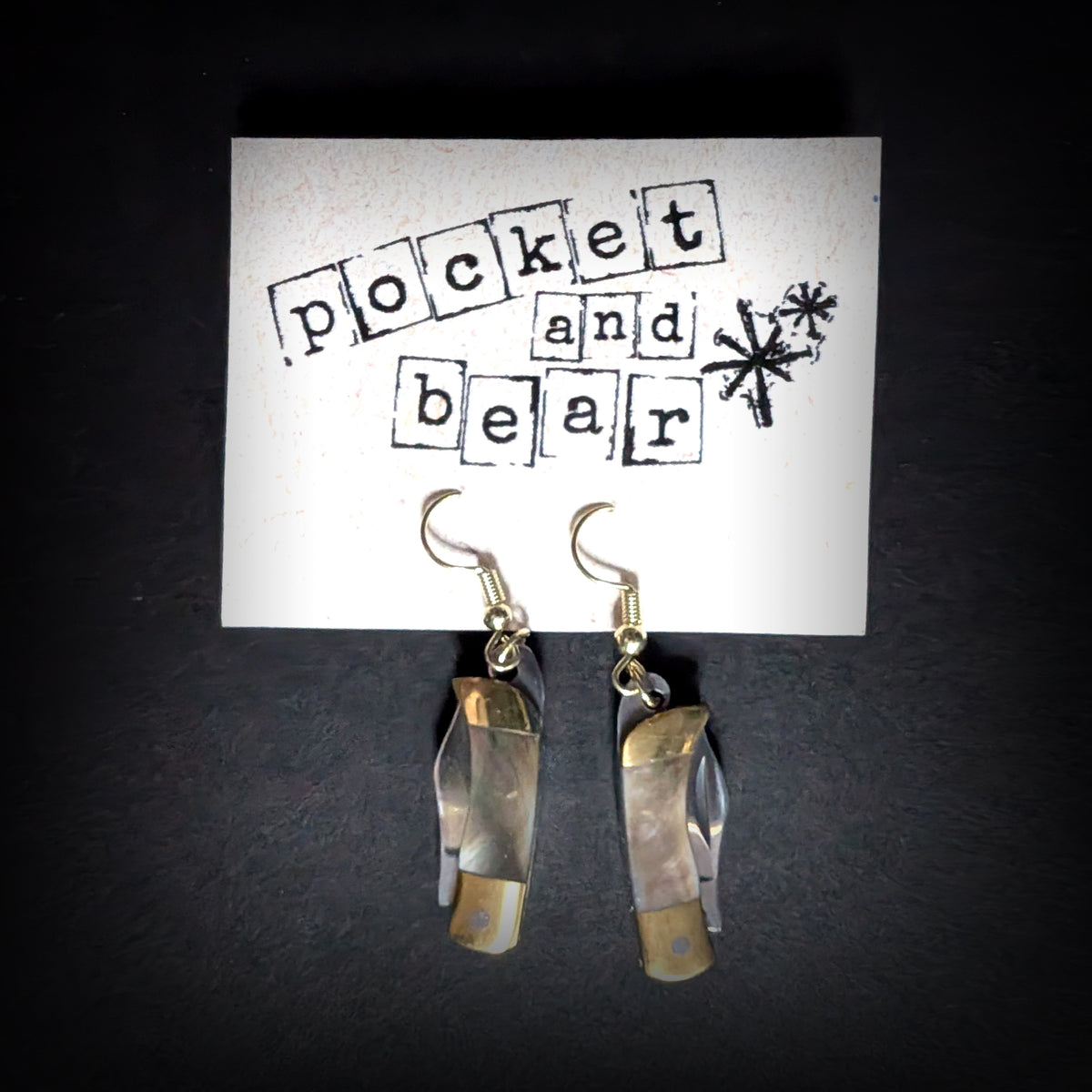 Dark shell mini pocket knife earrings by Pocket & Bear Collective hanging closed from a stamped Pocket and Bear card on grey, showing the smoky shell handles with brass caps