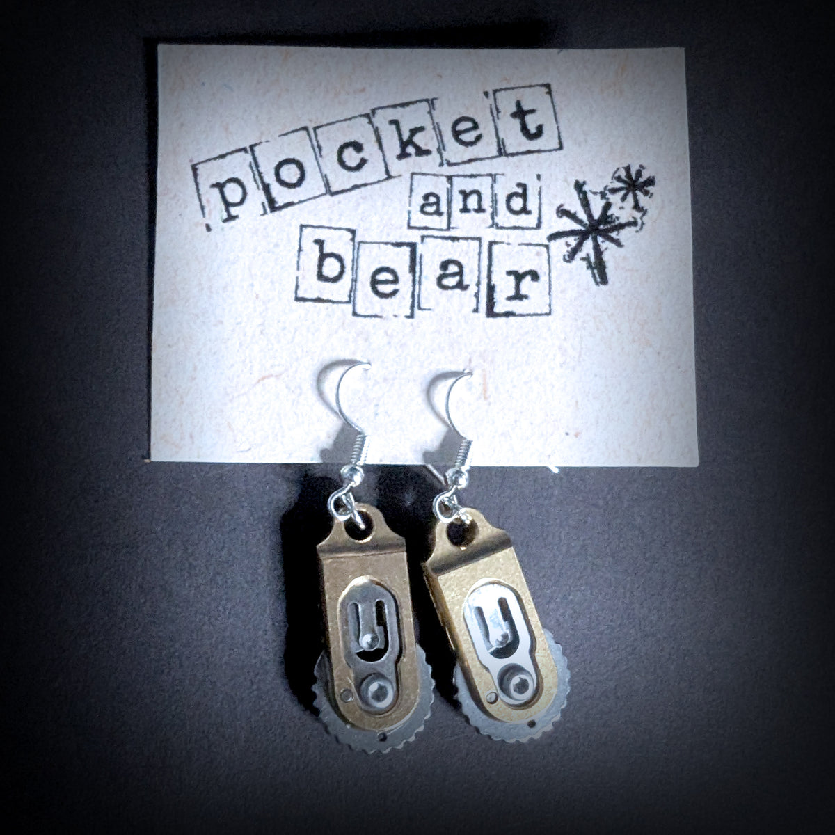 Gear pocket knife earrings by Pocket & Bear Collective hanging closed from a stamped Pocket and Bear card on a dark background