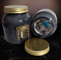 Two dark gray Just Breath spell candles in glass jars with gold lids and black and gold labels, one upright and one on its side showing white wax with dried lavender and a blue crescent moon stone, on black marble