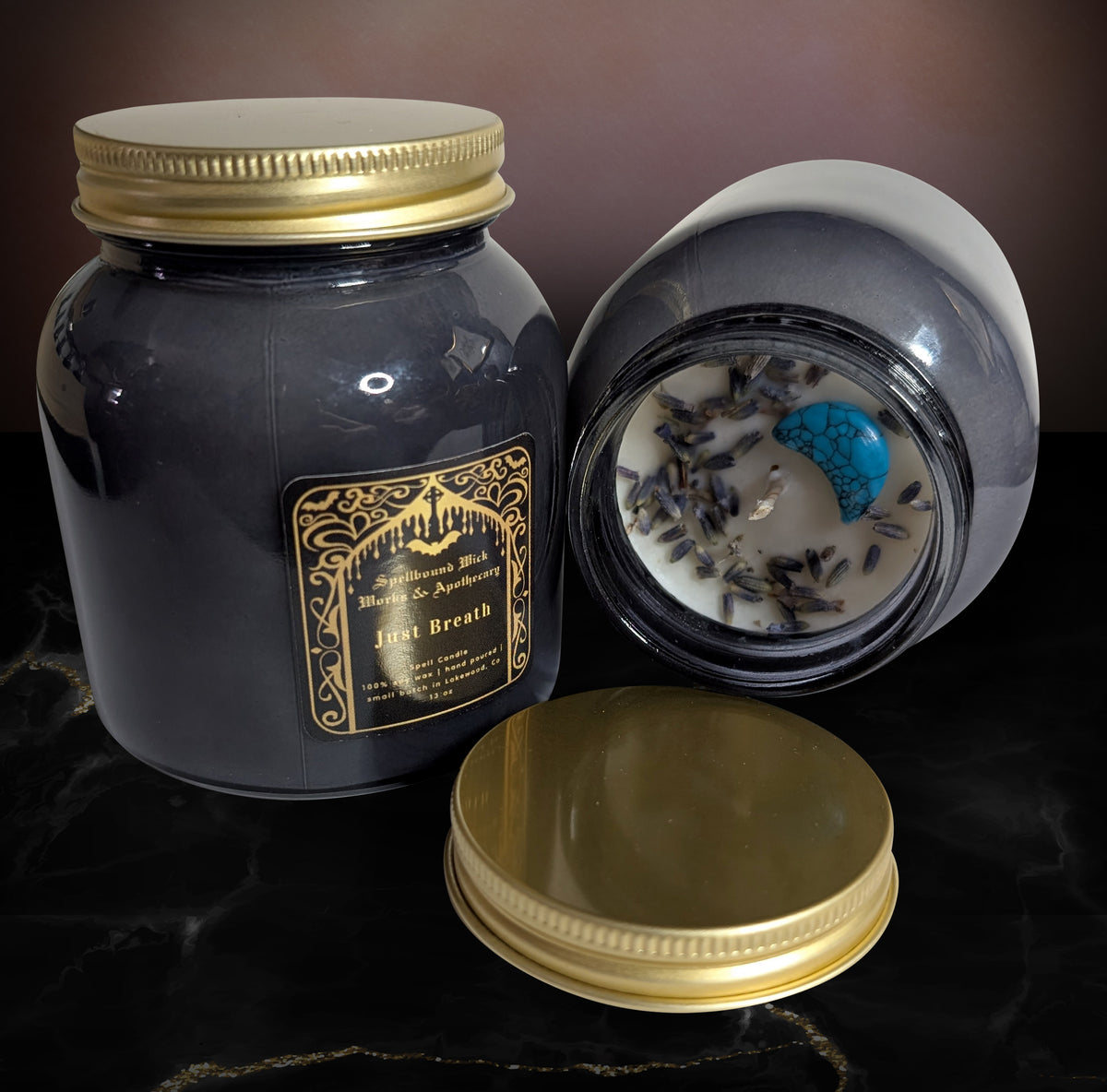 Two dark gray Just Breath spell candles in glass jars with gold lids and black and gold labels, one upright and one on its side showing white wax with dried lavender and a blue crescent moon stone, on black marble