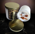 Persephone Goddess Candle by Spellbound Wick Works & Apothecary, a black and gold labelled soy candle with its gold lid off, beside a white dish holding a triple moon charm, dried botanicals and a brown stone on dark marble