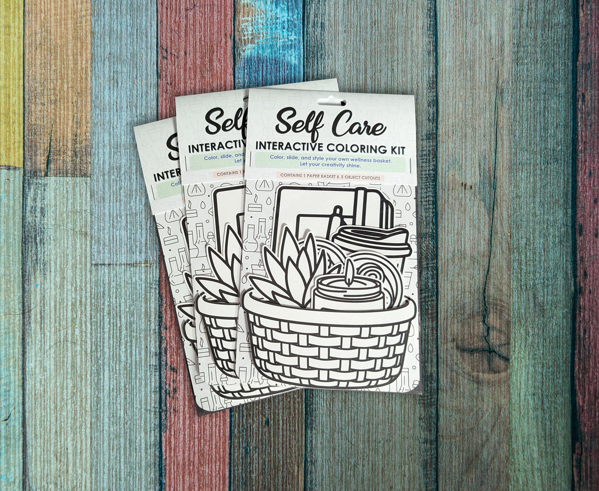 Three Self Care interactive coloring kits arranged on painted wood, each in its packaging with the uncolored basket and cutouts visible.