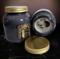 Black glass jar candle with a gold lid, shown open beside a second closed jar, the soy wax set with dark botanical pieces across the surface.