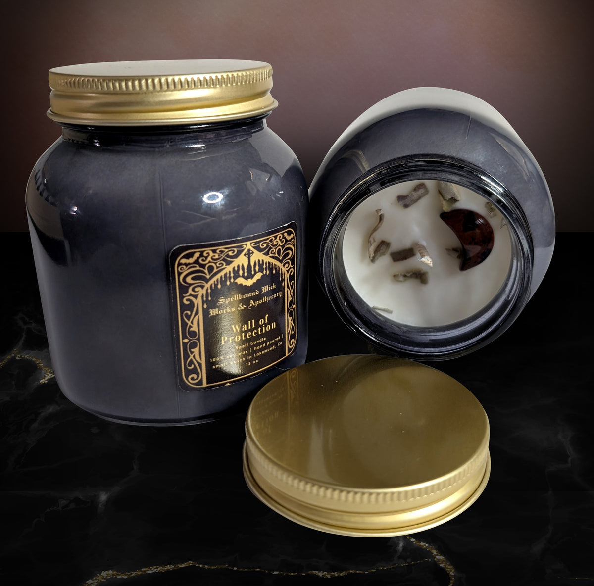 Black glass jar candle with a gold lid, shown open beside a second closed jar, the soy wax set with dark botanical pieces across the surface.