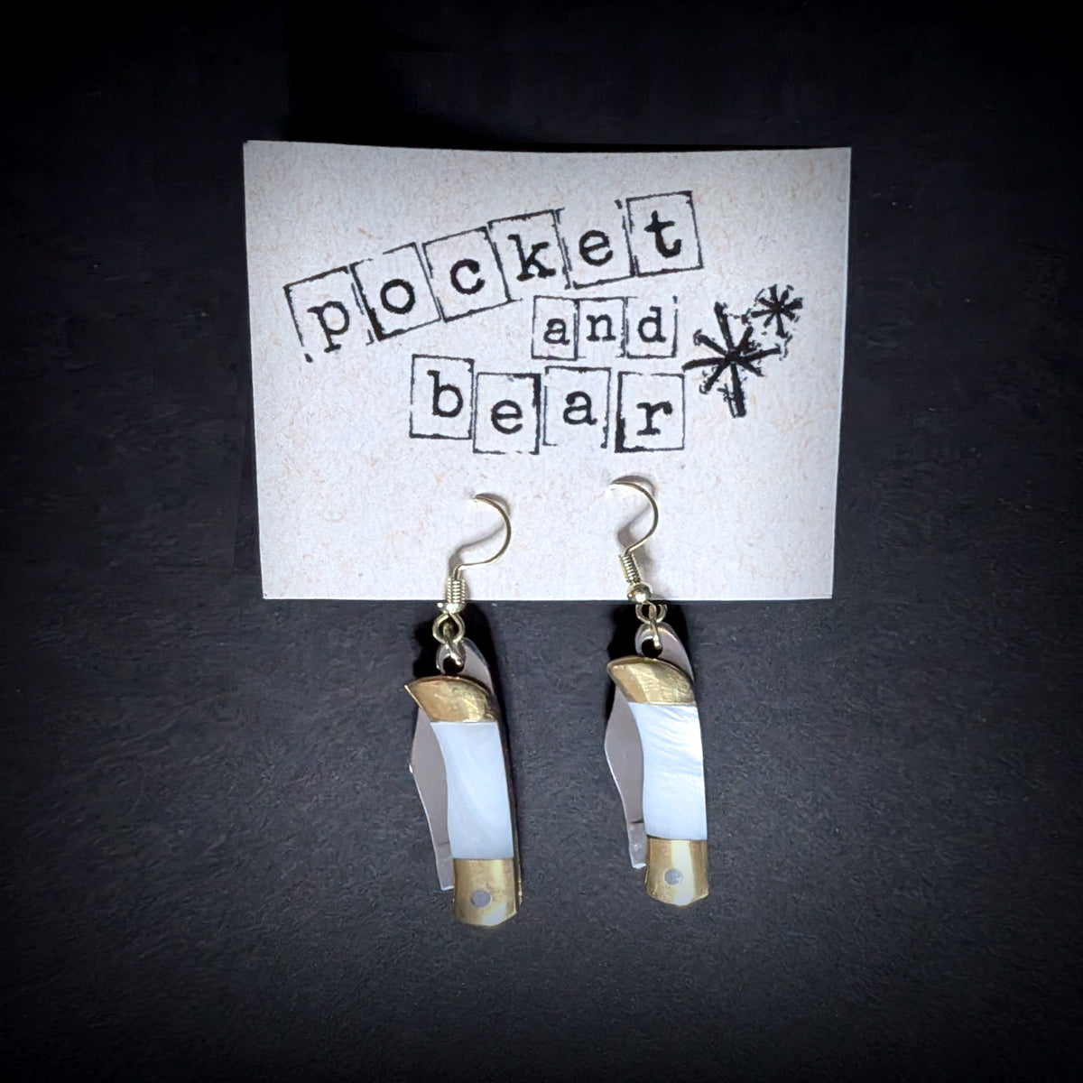 White shell mini pocket knife earrings by Pocket & Bear Collective hanging closed from a stamped Pocket and Bear card on grey, showing the white shell handles with brass caps