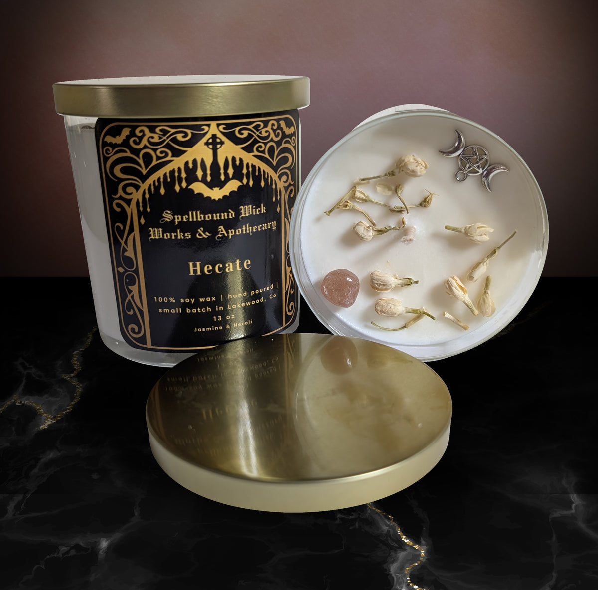Hecate Goddess Candle by Spellbound Wick Works & Apothecary, a black and gold labelled soy candle with its gold lid off, beside a white dish of small bone charms and a pink stone on dark marble