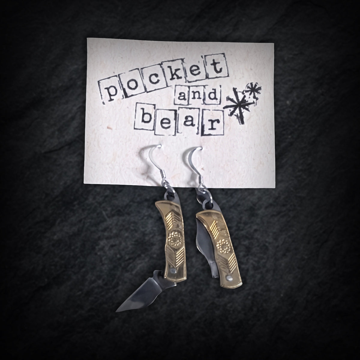 Brass flower mini pocket knife earrings by Pocket & Bear Collective hanging from a stamped Pocket and Bear card on dark slate, the left blade swung open and the right one closed, showing the engraved brass flower handles
