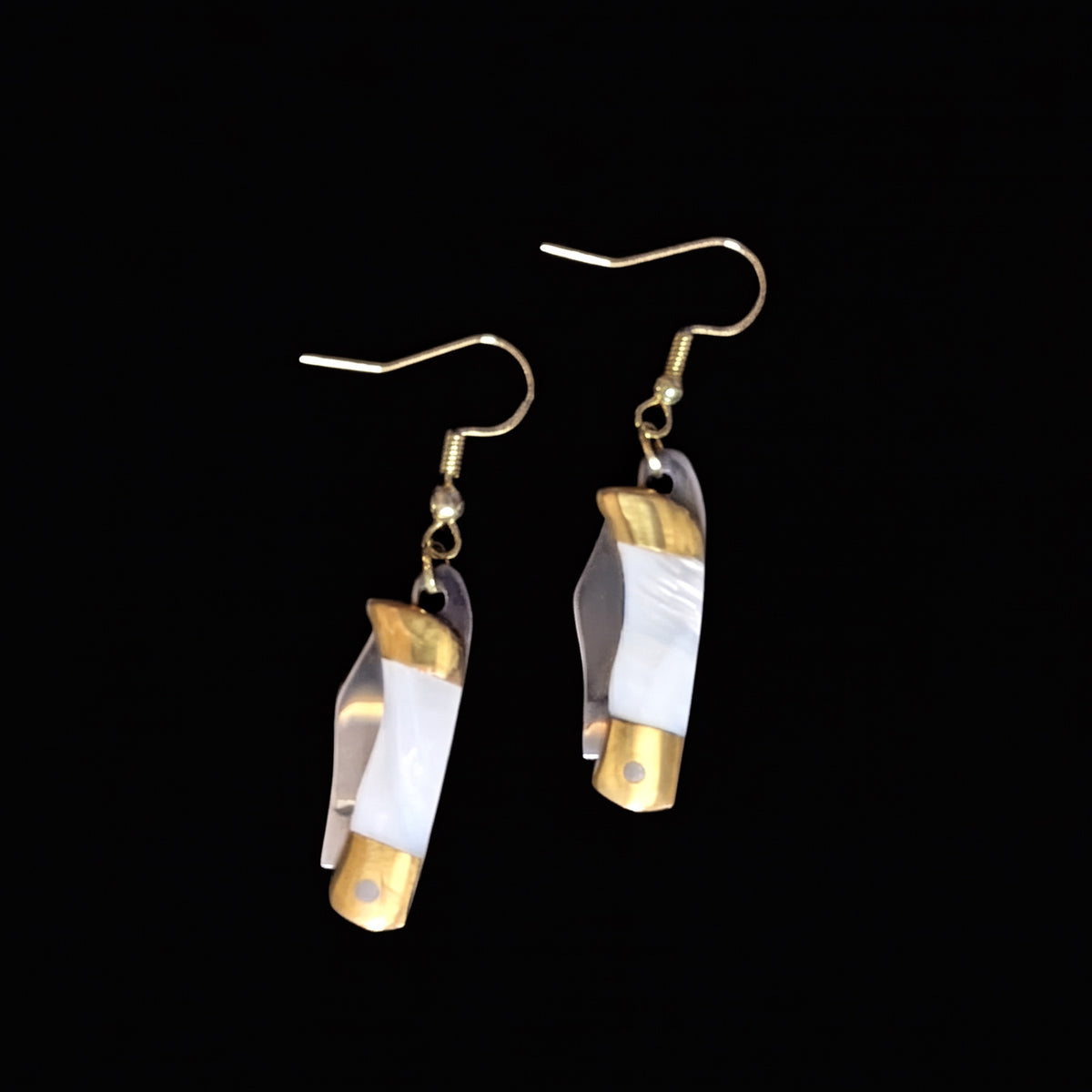 White shell mini pocket knife earrings by Pocket & Bear Collective closed, shown on their own against a black background with the gold plated earring hooks visible