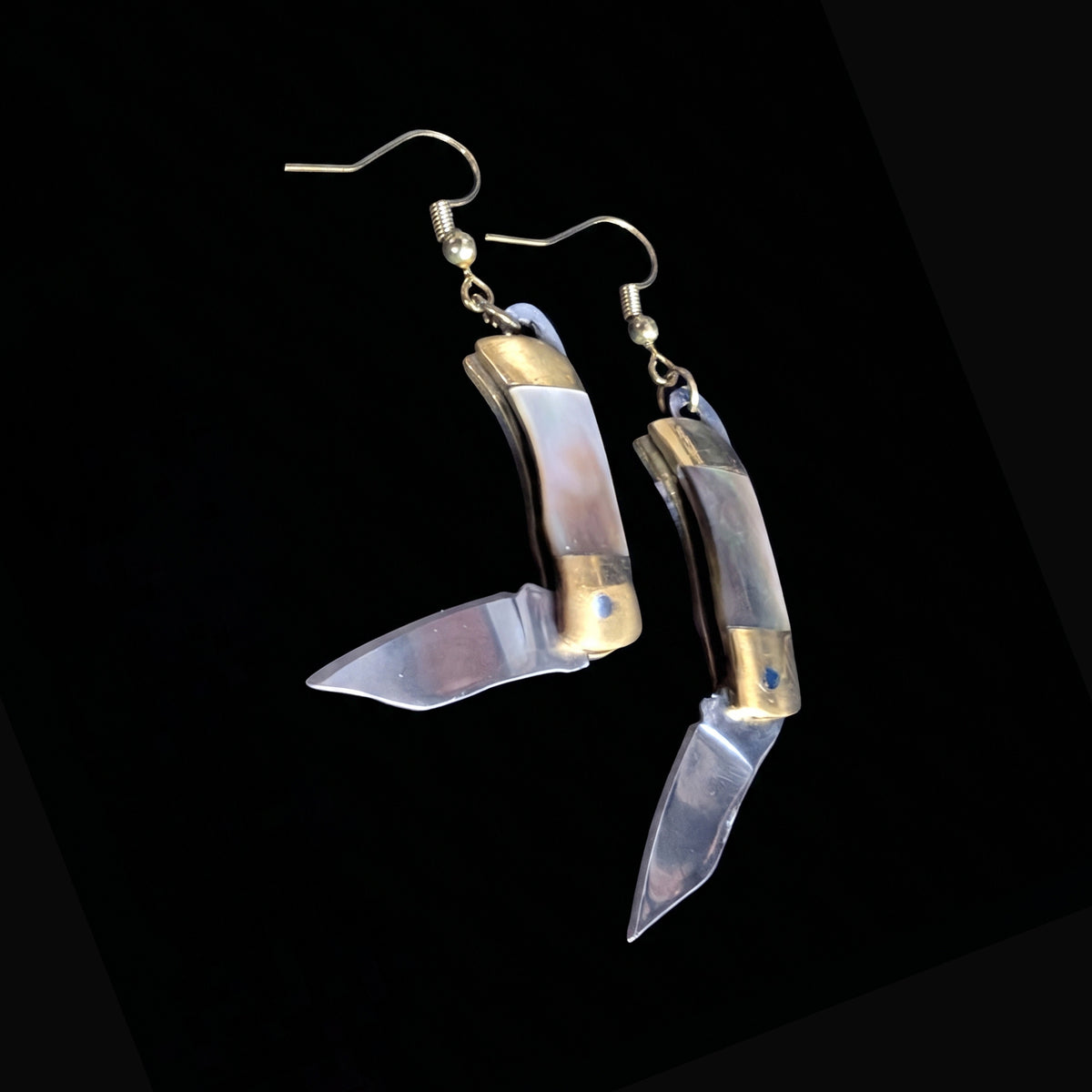 Dark shell mini pocket knife earrings by Pocket & Bear Collective on a black background with both stainless steel blades swung open, showing the pale shell handles and brass bolsters\