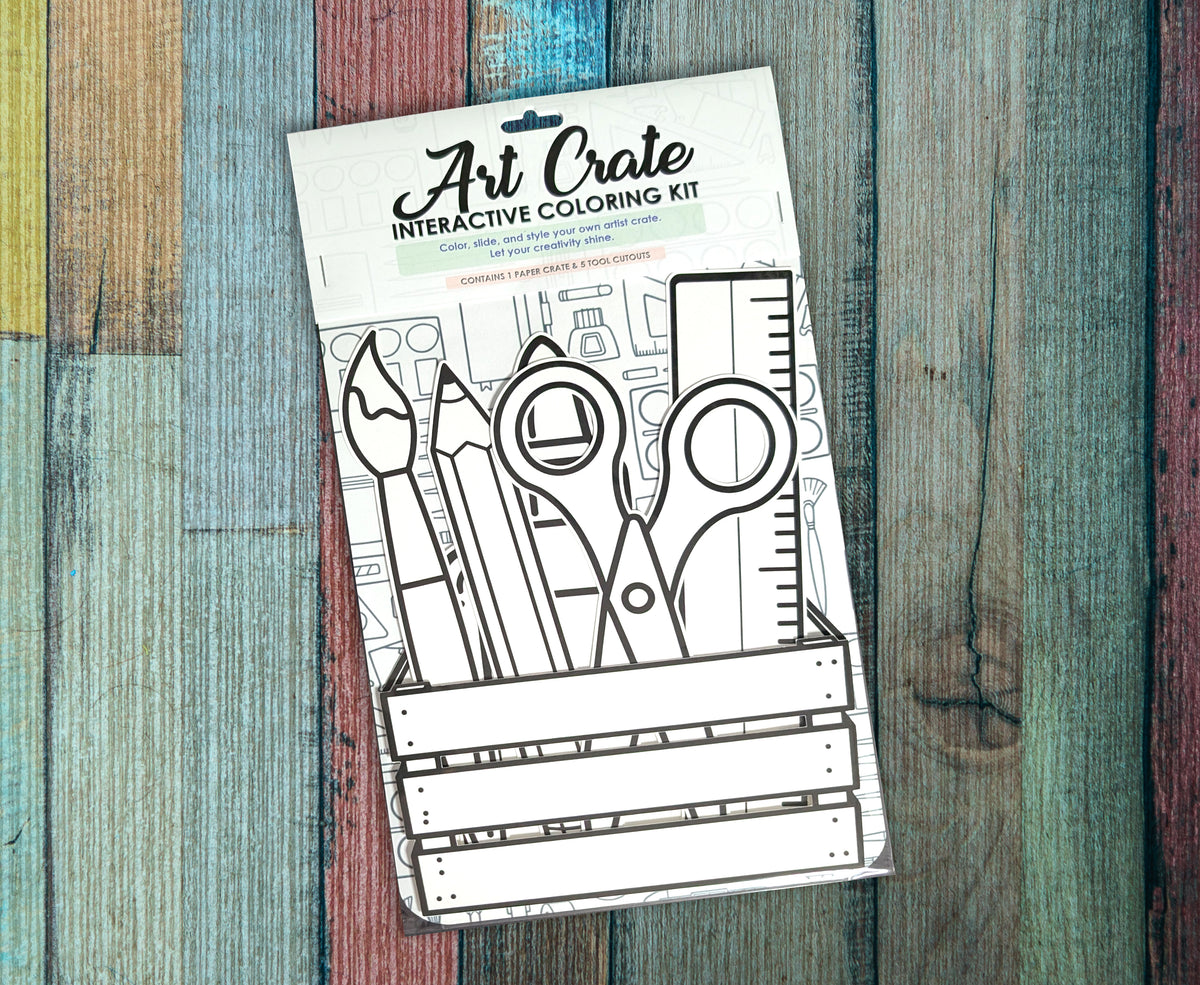 Single Art Crate interactive coloring kit lying on a painted wood background, the uncolored paintbrush, pencil, scissors and ruler cutouts showing through the front of the package below the Art Crate header card.