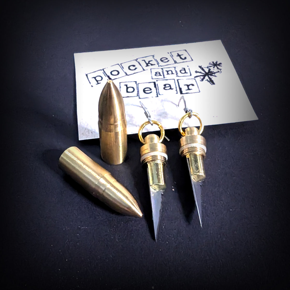 Bullet mini knife earrings by Pocket & Bear Collective, a pair of brass bullet shell earrings with the stainless steel blades open, angled on a stamped Pocket and Bear card beside a large brass bullet on a dark background
