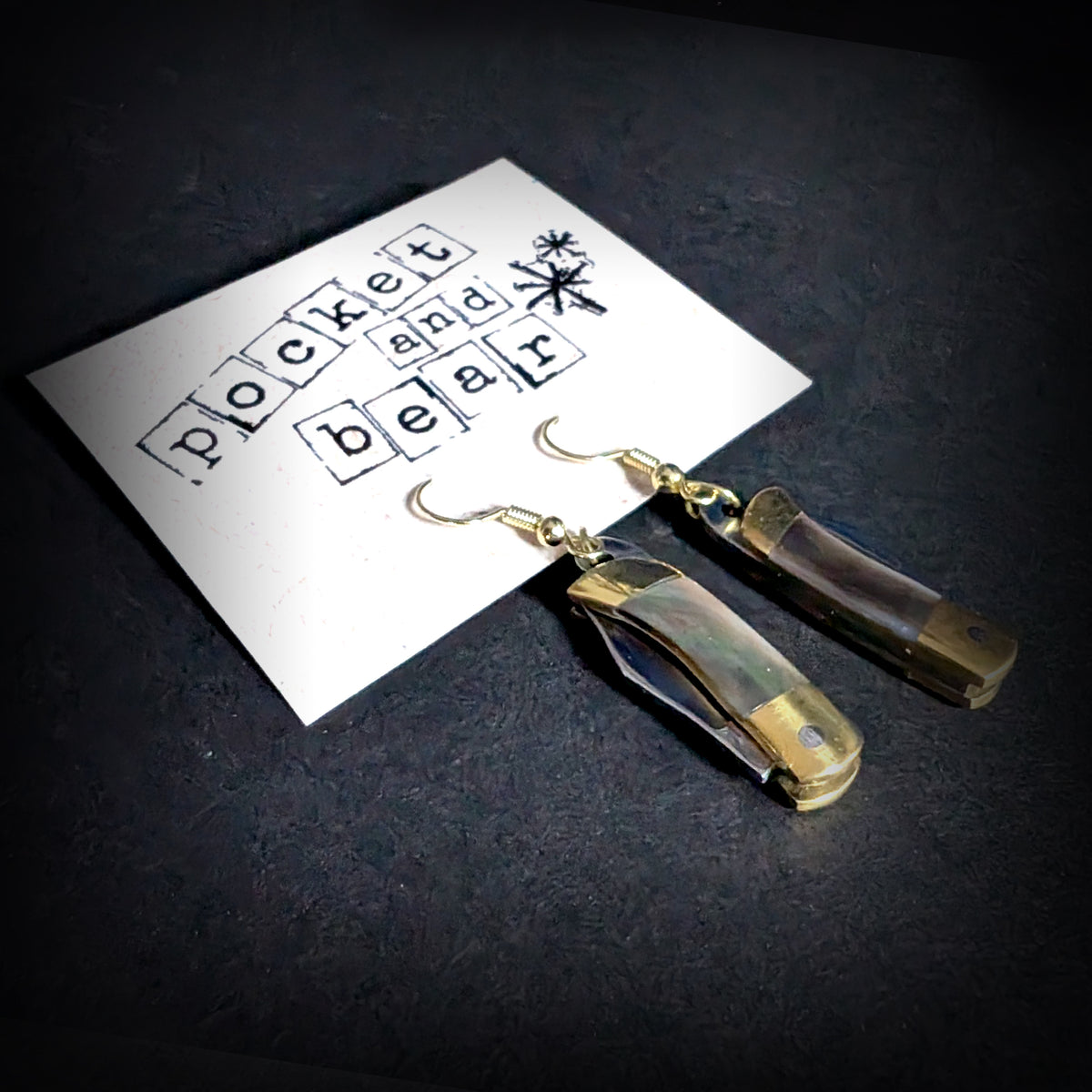 Dark shell mini pocket knife earrings by Pocket & Bear Collective hanging closed from a stamped Pocket and Bear card, shown close up at an angle