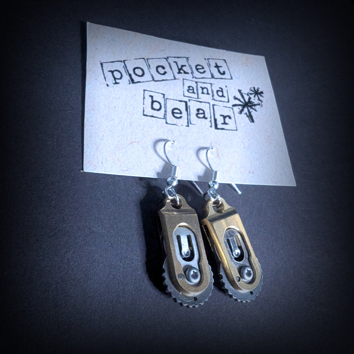 Gear pocket knife earrings by Pocket & Bear Collective hanging closed from a stamped Pocket and Bear card, shown close up, with the gear-toothed brass handles in view