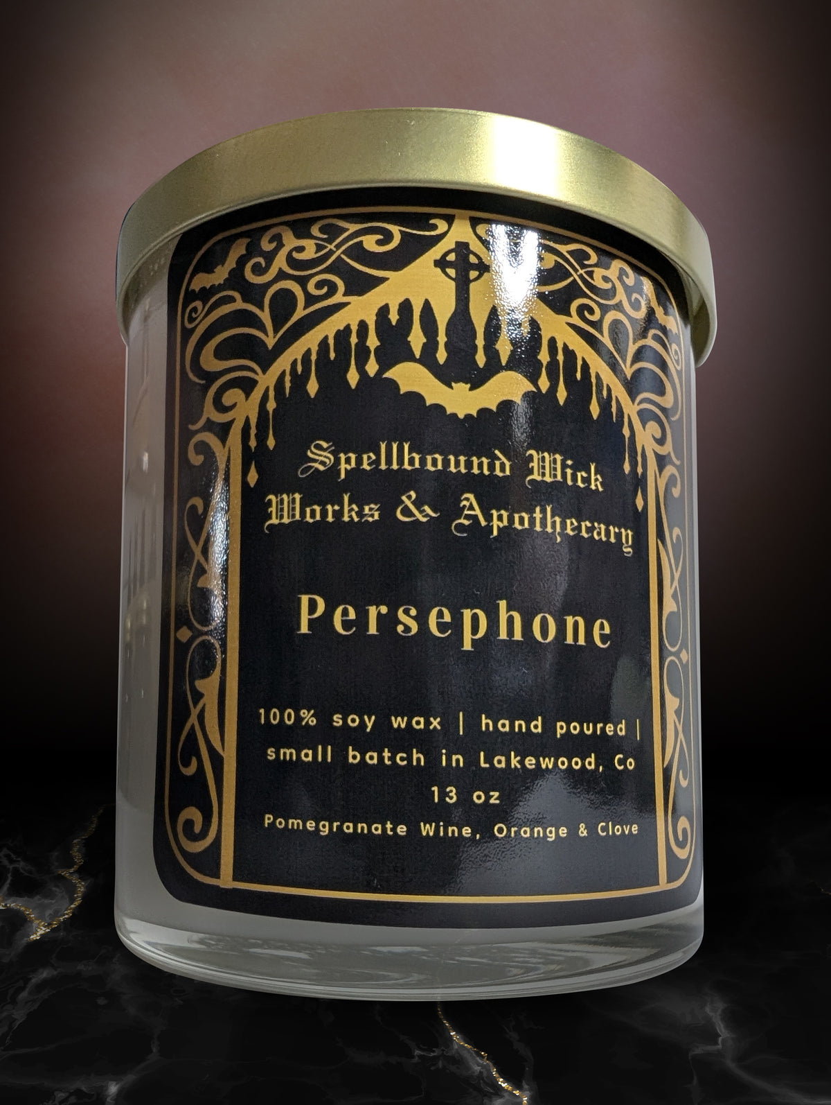 The Persephone Goddess Candle closed with its gold lid on, label facing forward on dark marble