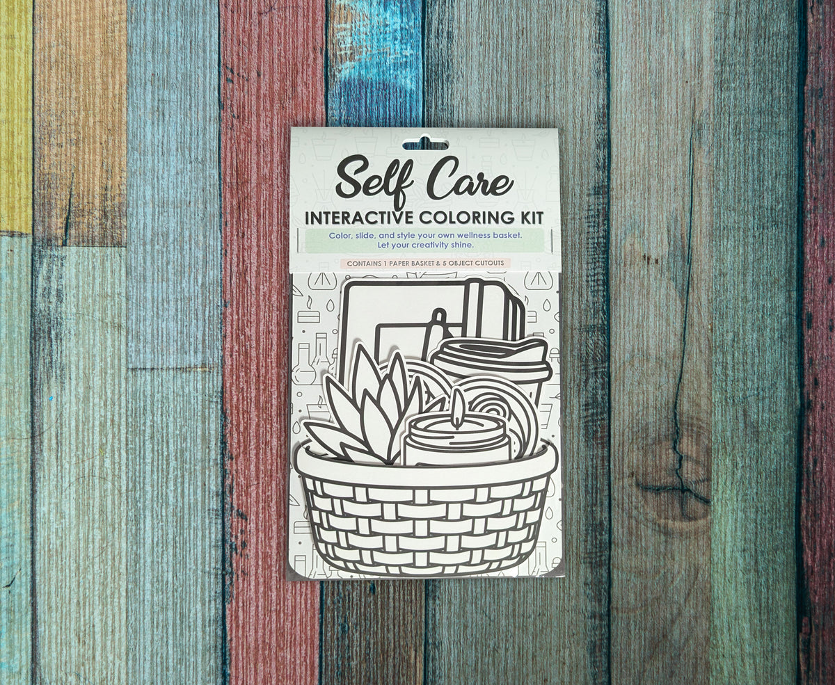 Single Self Care interactive coloring kit lying on a painted wood background, the uncolored basket and cutouts showing through the front of the package below the Self Care header card.