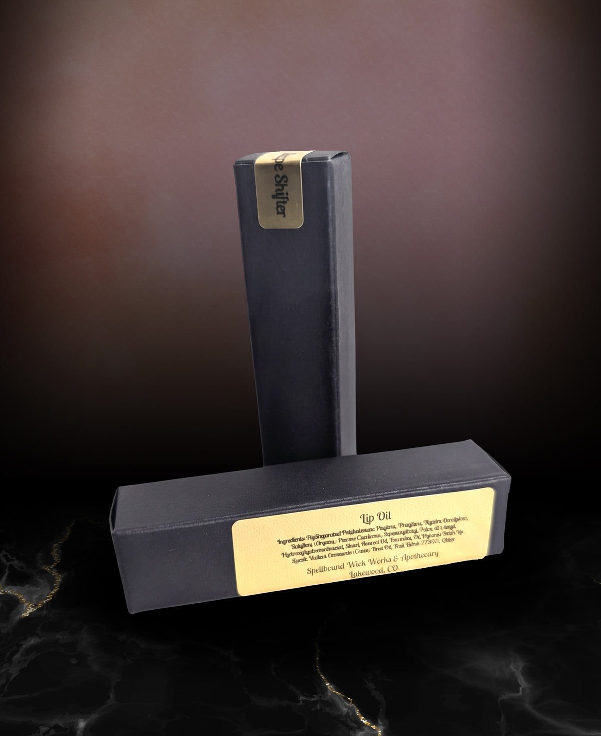 Closed black lip oil bottle standing upright on dark marble beside its black box, the gold label reading Lip Oil by Spellbound Wick Works & Apothecary.