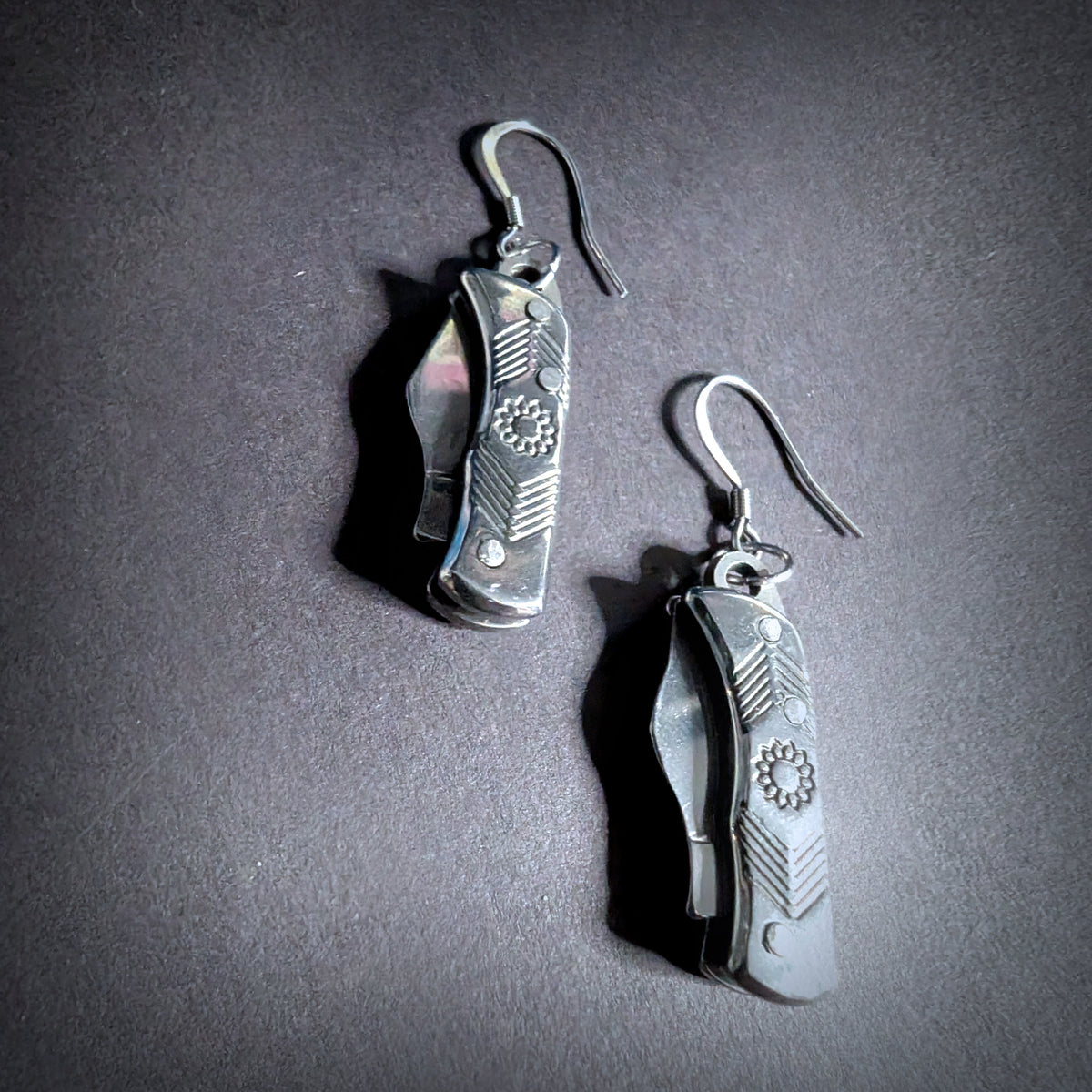 Silver flower mini pocket knife earrings by Pocket & Bear Collective, the pair closed on dark grey, shown close up with the engraved flower and line work on the stainless steel handles