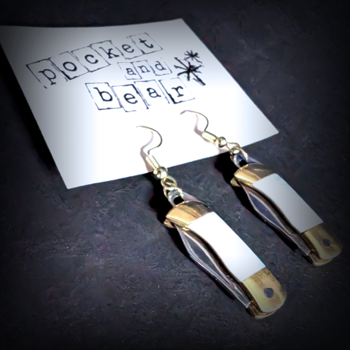 White shell mini pocket knife earrings by Pocket & Bear Collective hanging closed from a stamped Pocket and Bear card, shown close up at an angle