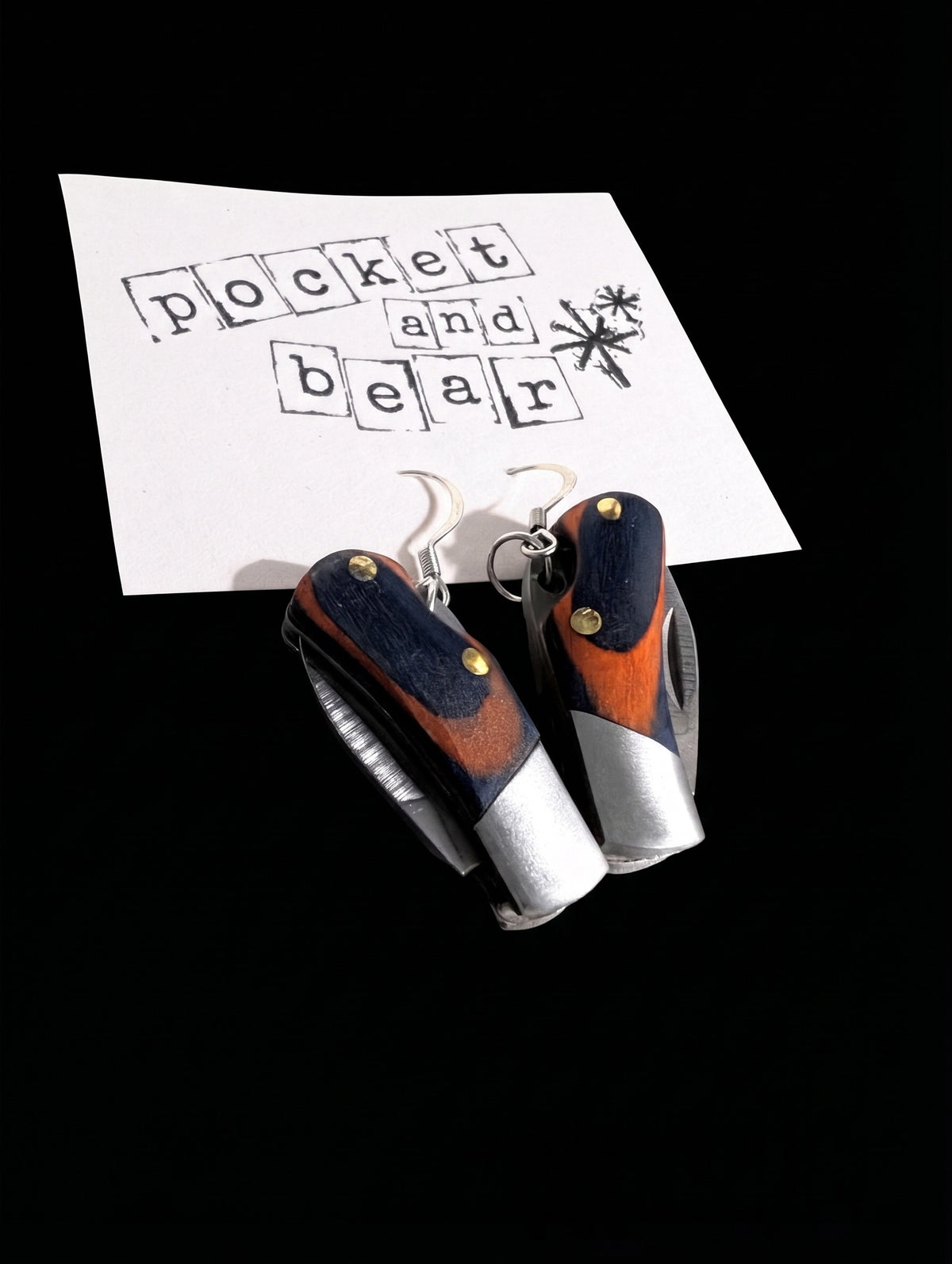Wooden mini pocket knife earrings by Pocket & Bear Collective, the pair hanging closed from a stamped Pocket and Bear card on a grey background, showing the orange and dark blue wood handles