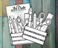 Two Art Crate interactive coloring kits in their packaging on a painted wood background, showing the uncolored paintbrush, pencil, scissors and ruler cutouts above the crate.