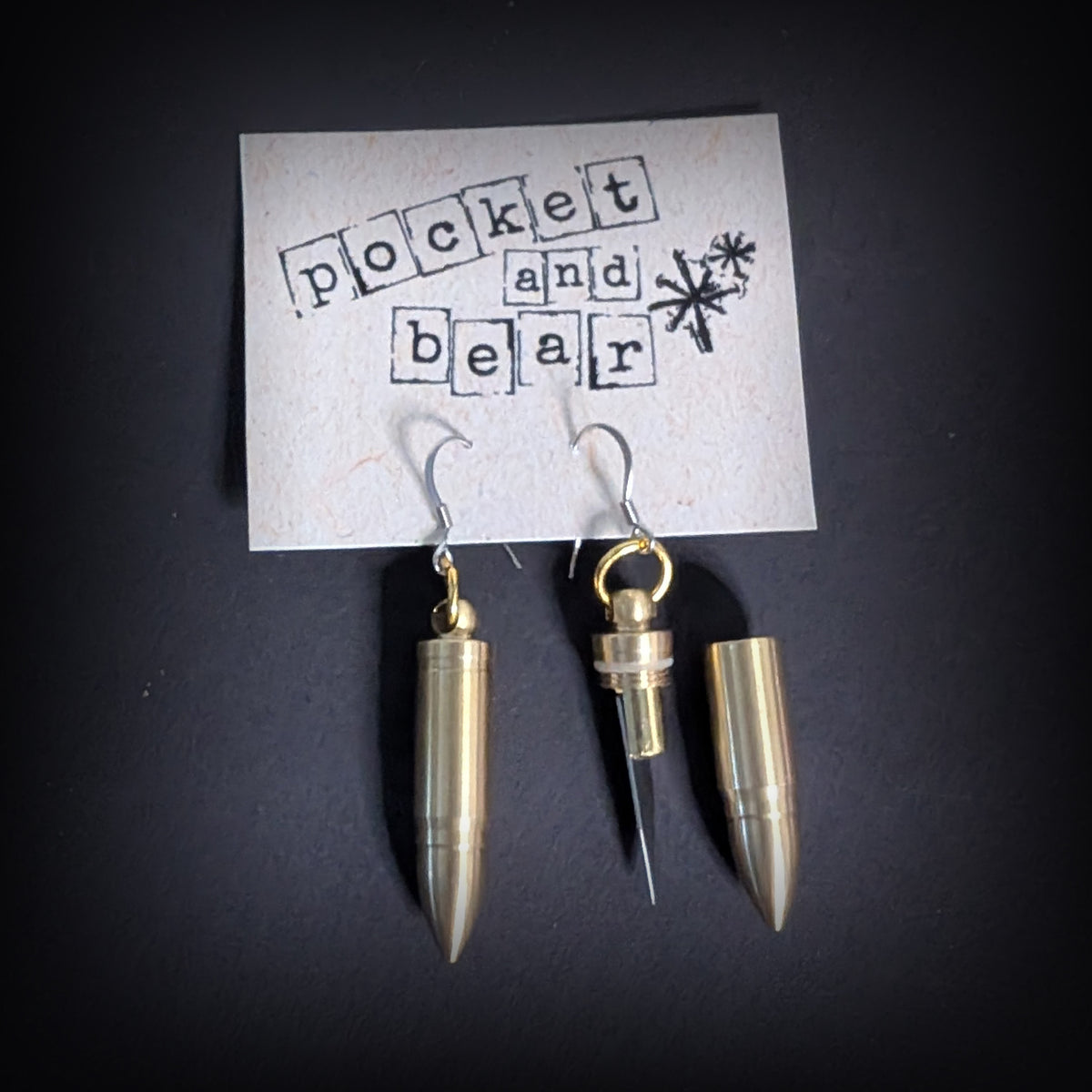 Bullet mini knife earrings by Pocket & Bear Collective, the pair hanging from a stamped Pocket and Bear card with one blade open and one closed, on a dark background