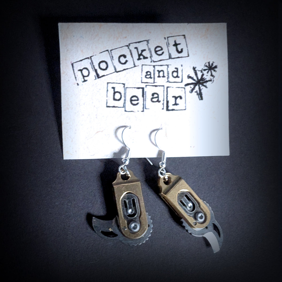 Gear pocket knife earrings by Pocket & Bear Collective hanging from a stamped Pocket and Bear card with both stainless steel blades swung open, shown close up