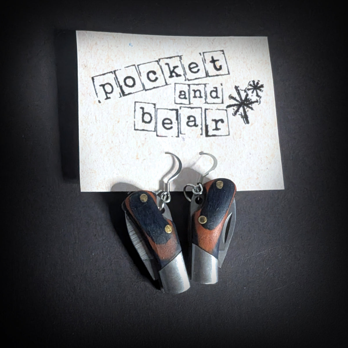 Wooden mini pocket knife earrings by Pocket & Bear Collective hanging closed from a stamped Pocket and Bear card on a black background, with the silver plated earring hooks visible