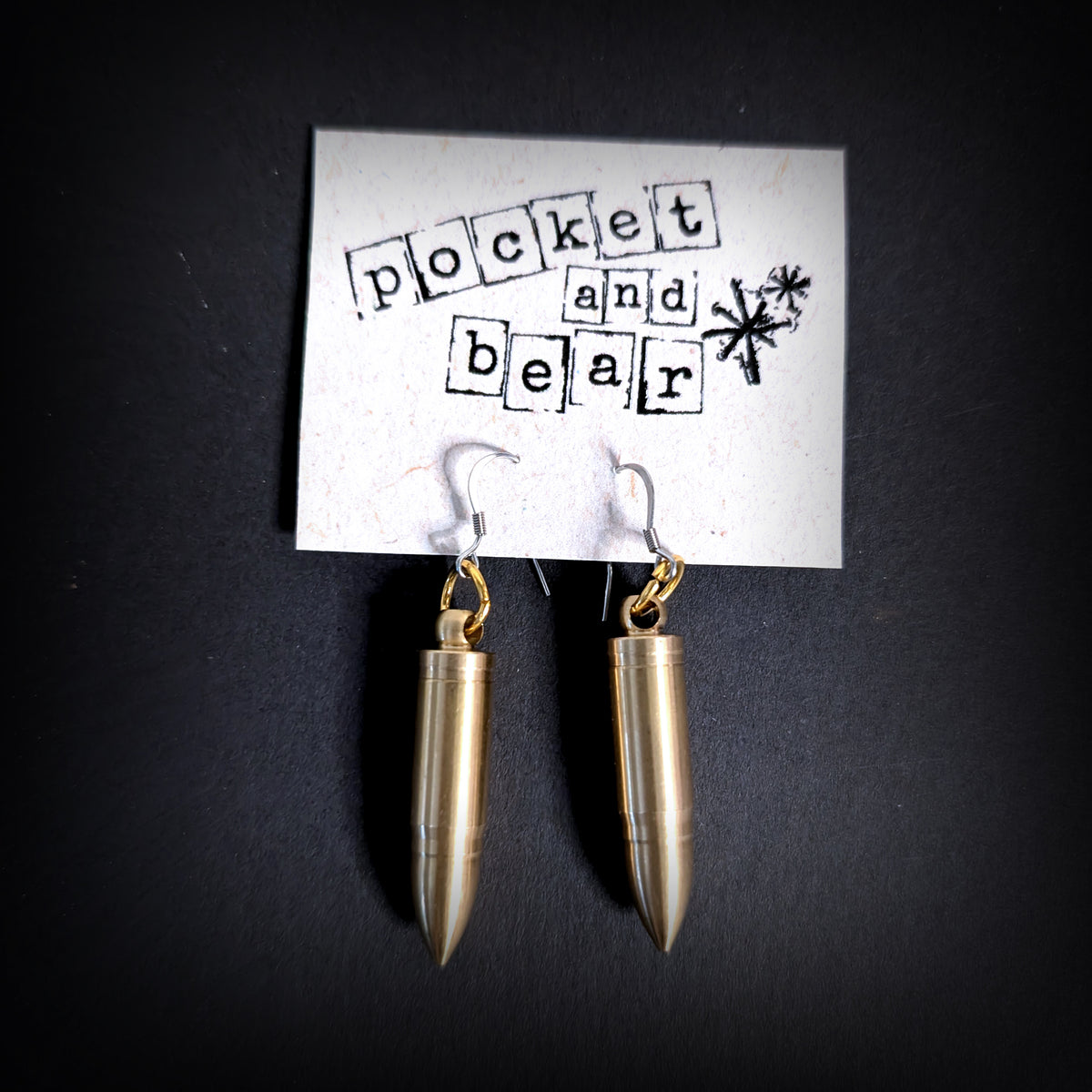Bullet mini knife earrings by Pocket & Bear Collective, both blades closed inside the brass bullet shells, hanging from a stamped Pocket and Bear card on a dark background