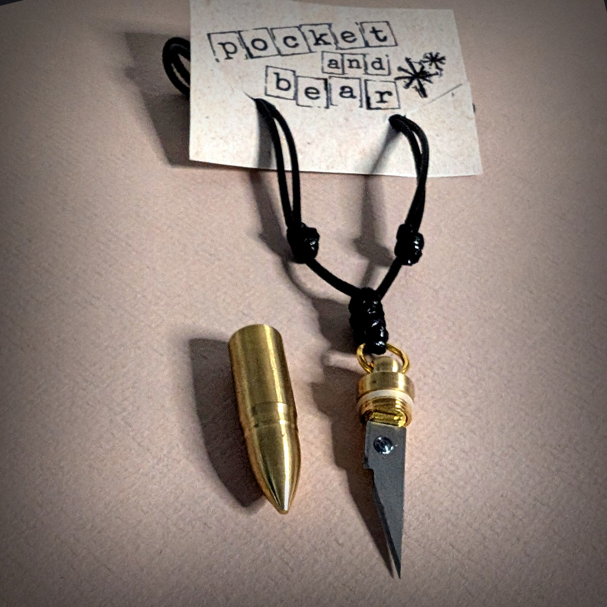 Bullet mini knife necklace by Pocket & Bear Collective laid flat on a cream background with a stamped Pocket and Bear card, the stainless steel blade pulled out of its brass bullet shell