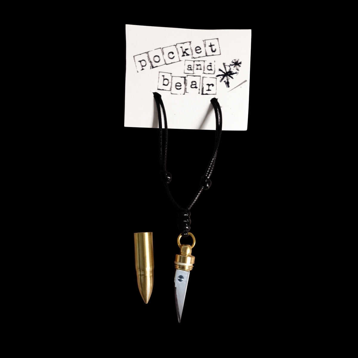 Bullet mini knife necklace by Pocket & Bear Collective hanging from a stamped Pocket and Bear card on a black background, the stainless steel blade pulled out and the empty brass bullet shell resting beside it