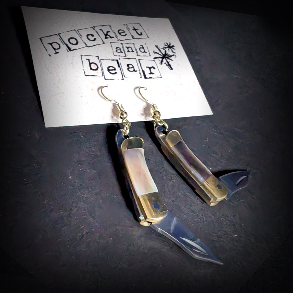 Dark shell mini pocket knife earrings by Pocket & Bear Collective hanging from a stamped Pocket and Bear card with one stainless steel blade swung open
