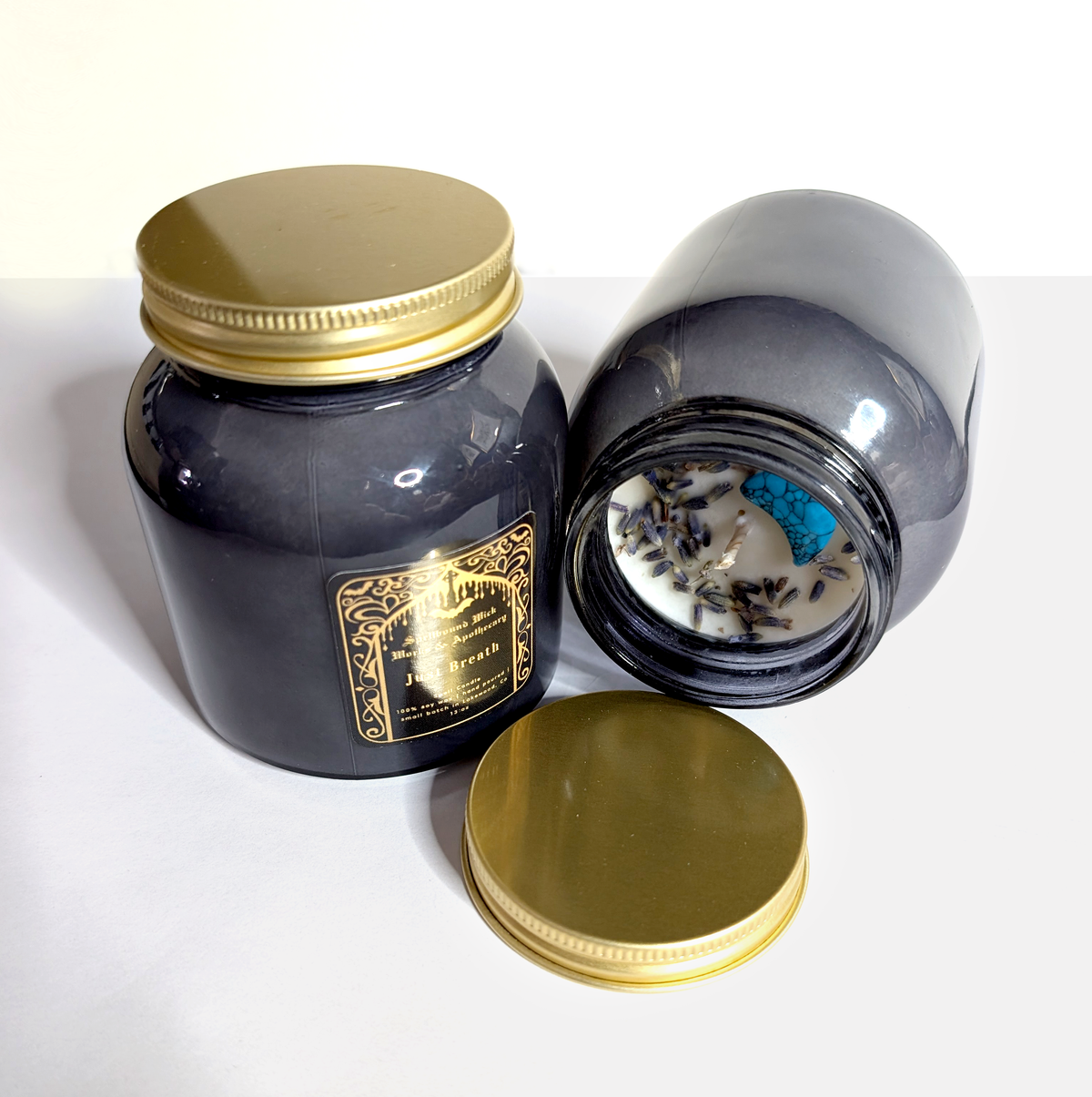Just Breath spell candle jar tilted to show the wax top with dried lavender and a blue stone, gold lid resting beside it, on a white background