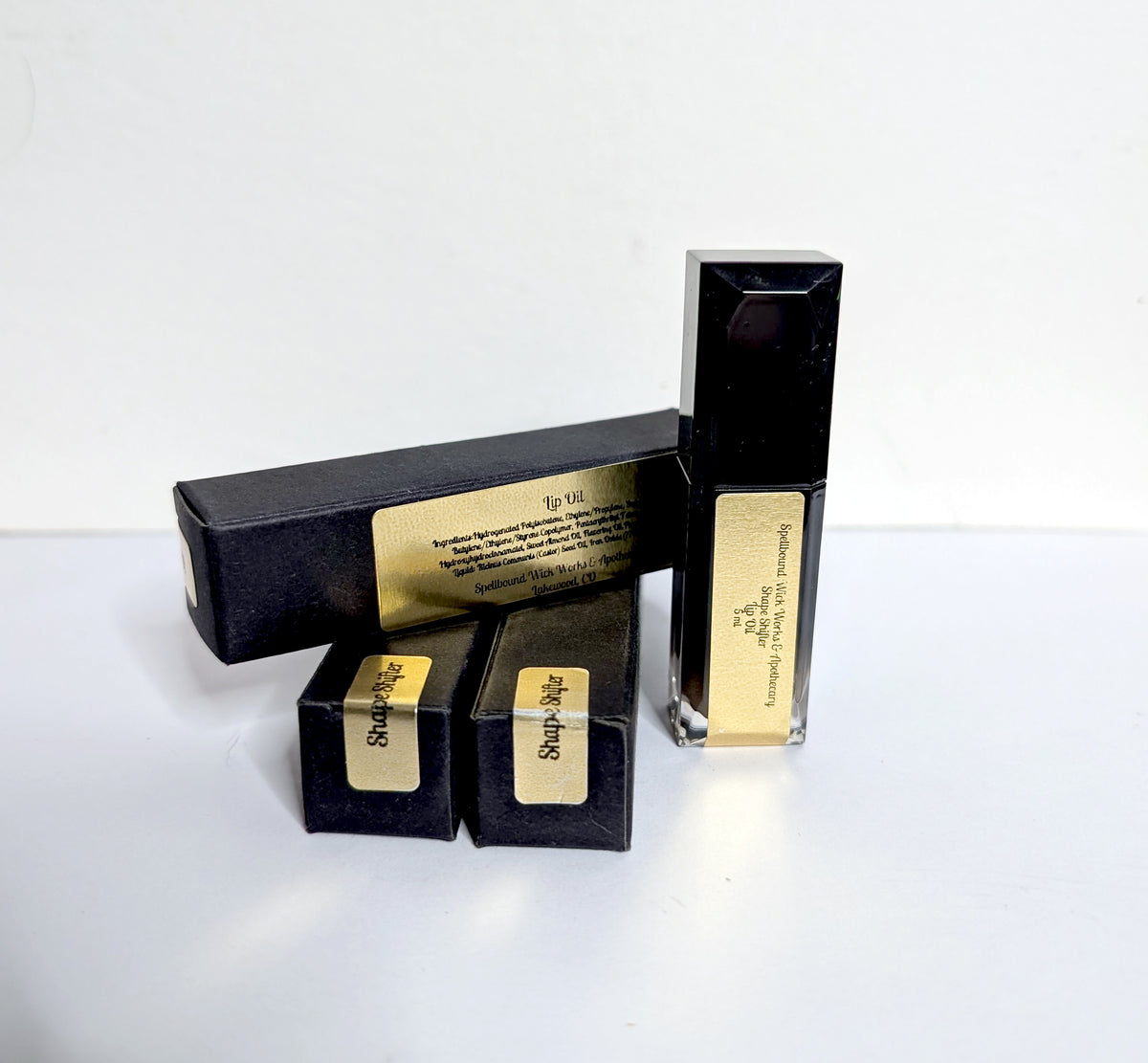 Black lip oil bottle and boxes photographed together against a pale background, gold labels facing the camera.