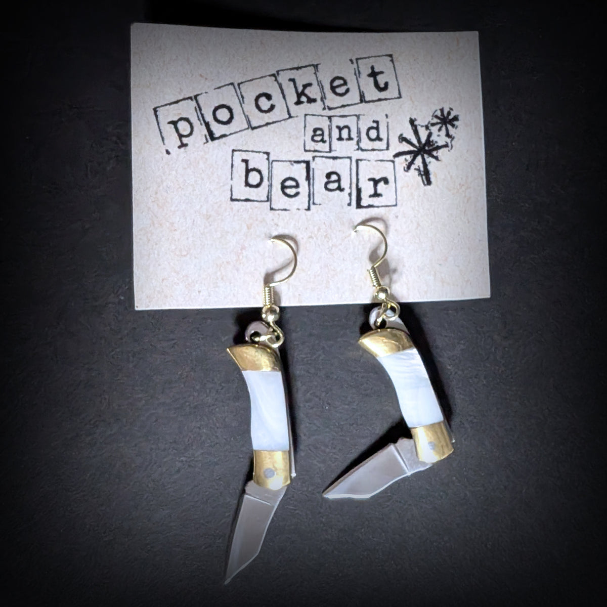 White shell mini pocket knife earrings by Pocket & Bear Collective hanging from a stamped Pocket and Bear card with one stainless steel blade swung open