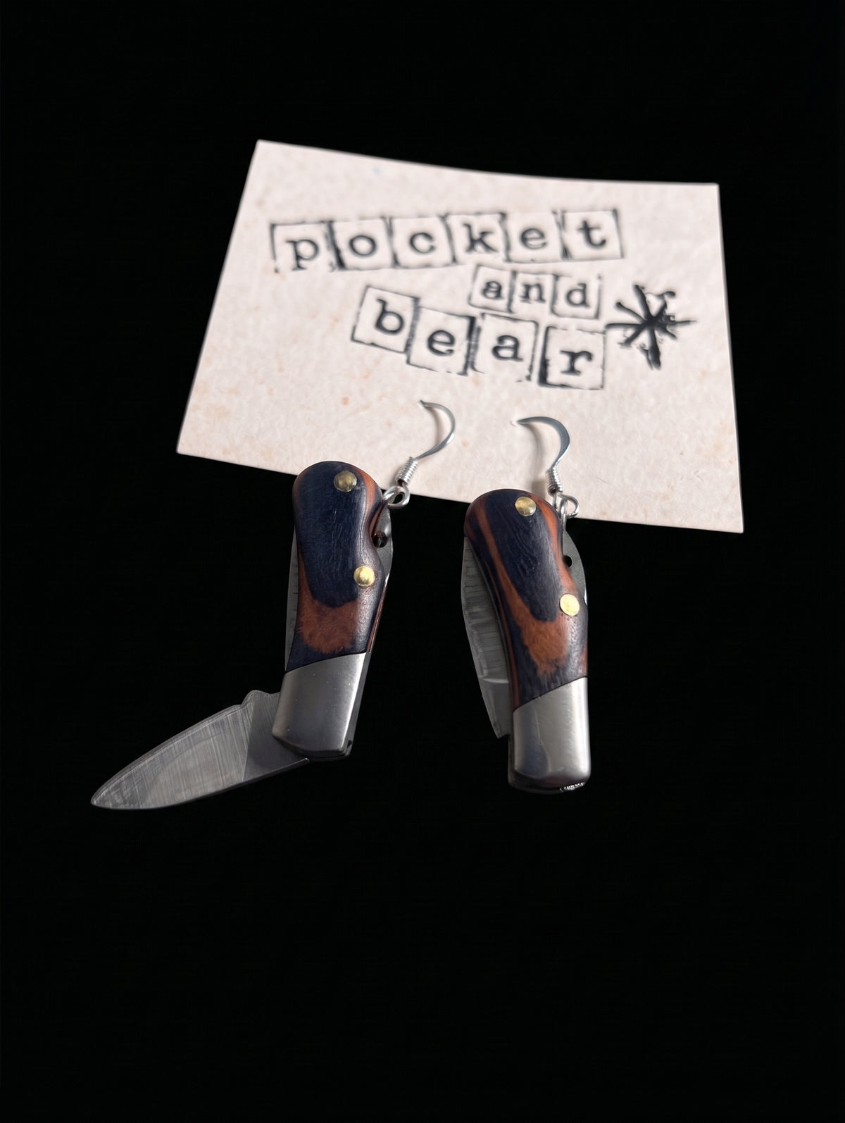 Wooden mini pocket knife earrings by Pocket & Bear Collective shown from overhead resting on a stamped Pocket and Bear card, both blades open