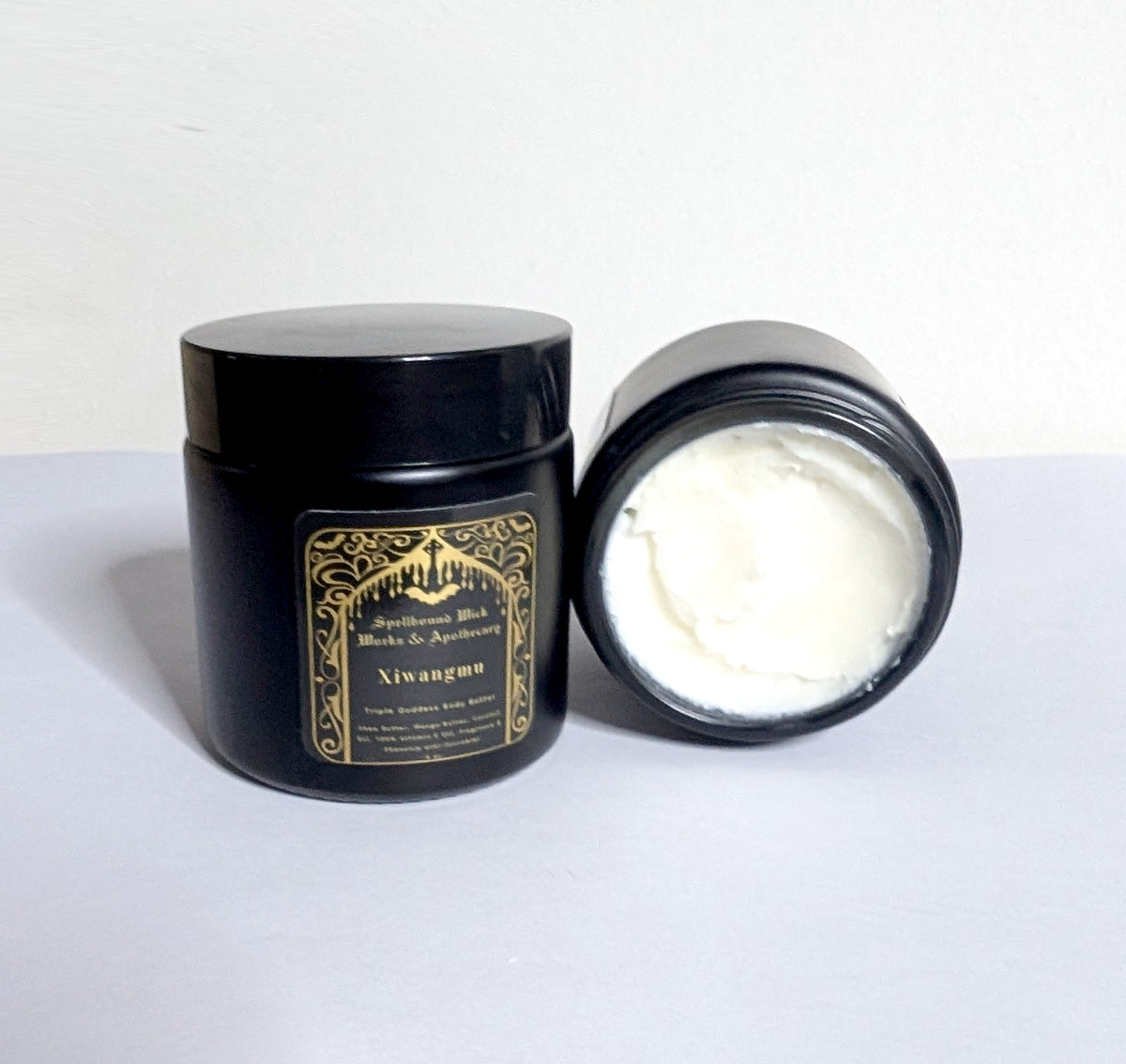 Open black jar showing the whipped white body butter, lid set behind it on a white background.