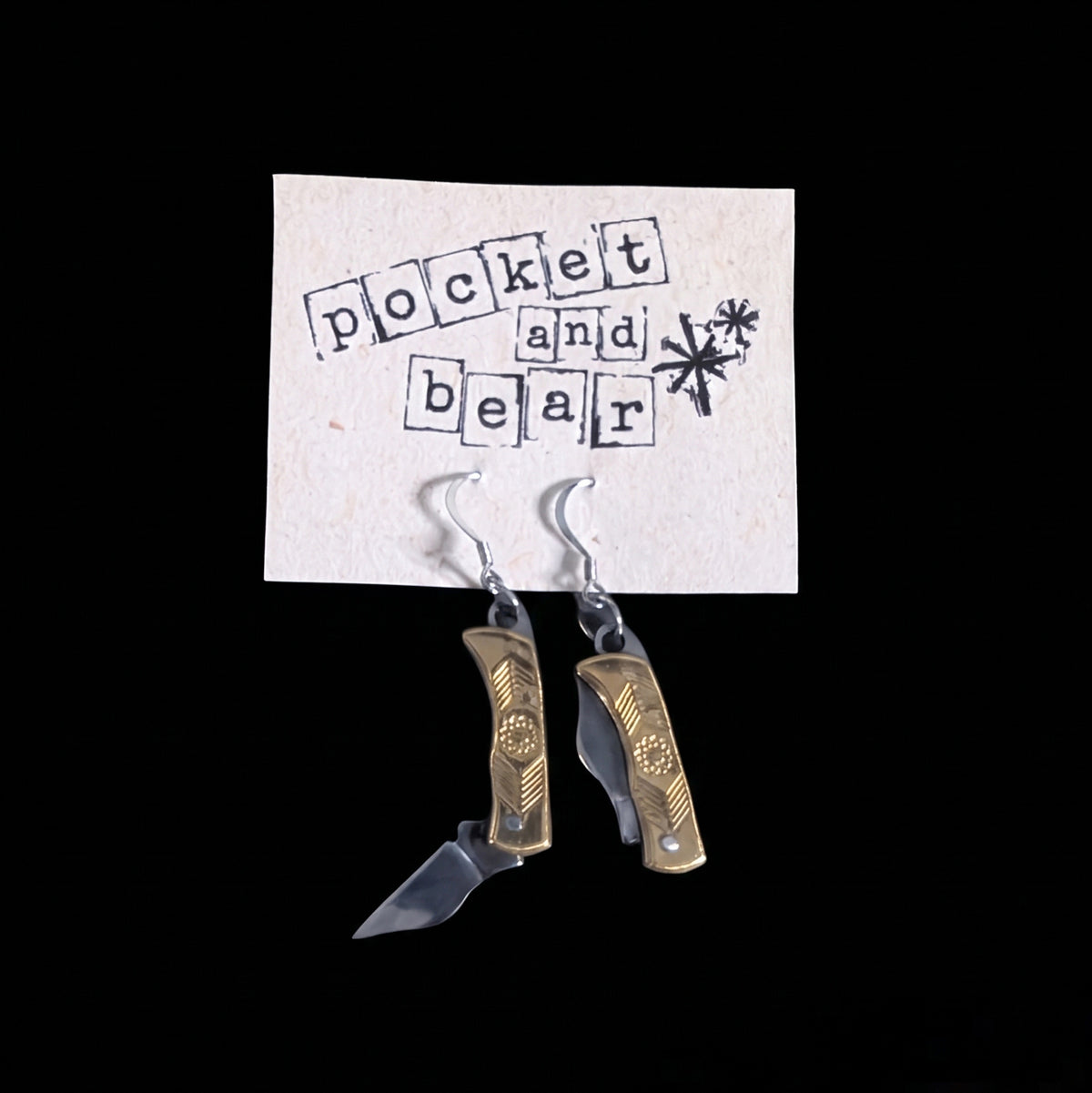 Brass flower mini pocket knife earrings by Pocket & Bear Collective hanging from a stamped Pocket and Bear card with one blade open, on a black background