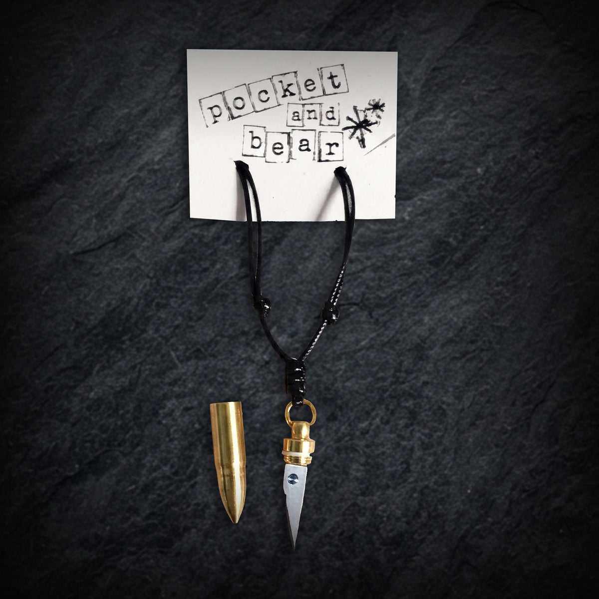 Bullet mini knife necklace by Pocket & Bear Collective hanging from a stamped Pocket and Bear card on dark slate, the stainless steel blade pulled out and the empty brass bullet shell resting beside it
