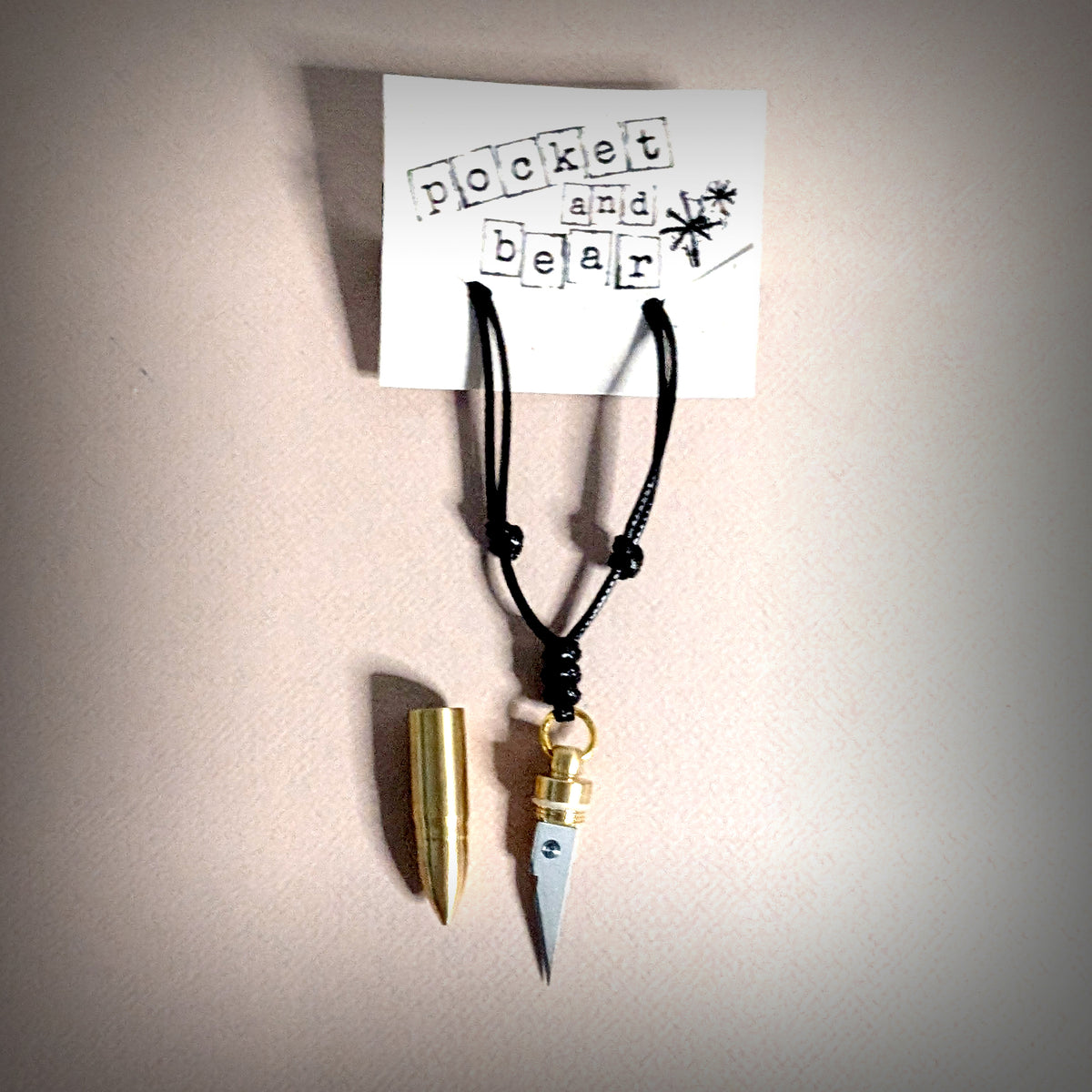 Bullet mini knife necklace by Pocket & Bear Collective shown from overhead on a cream background with a stamped Pocket and Bear card, the stainless steel blade pulled out of its brass bullet shell