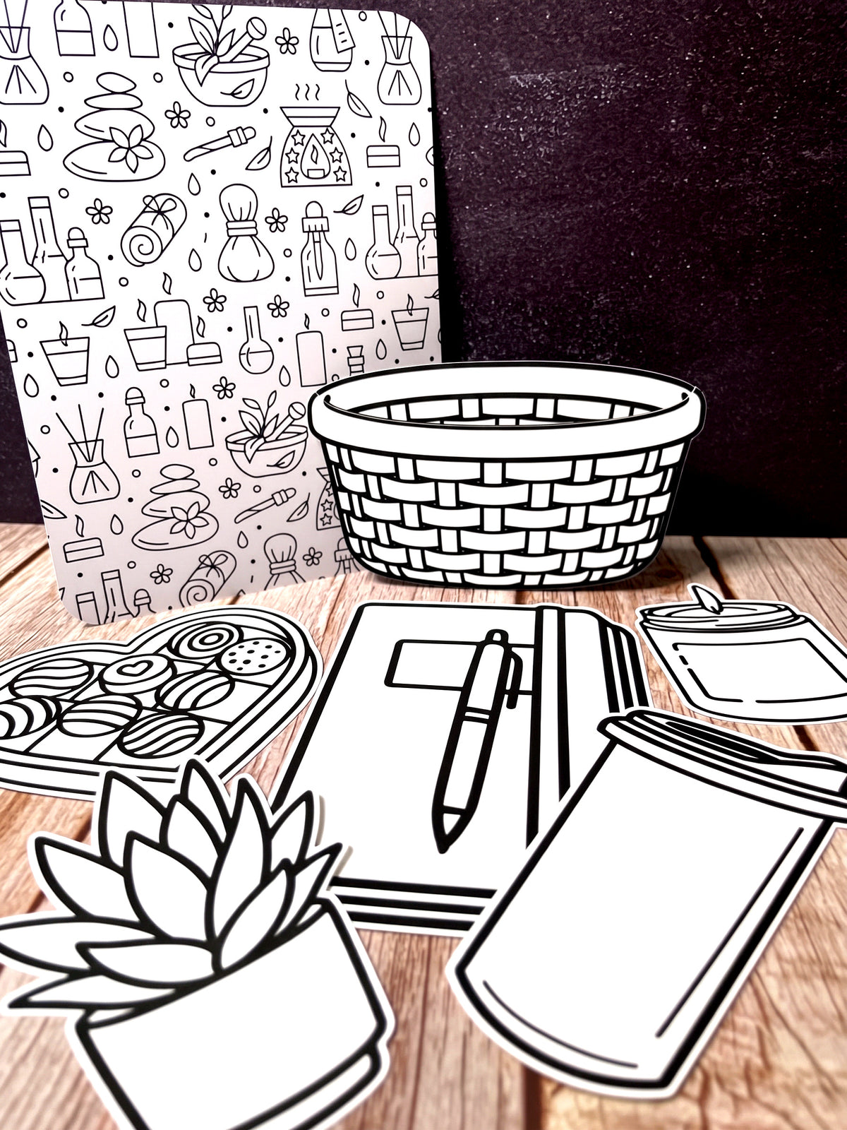 The five Self Care cutouts laid out on wood, the woven basket standing in front of the art board with a potted succulent, a journal and pen, a box of chocolates and a candle arranged below it.