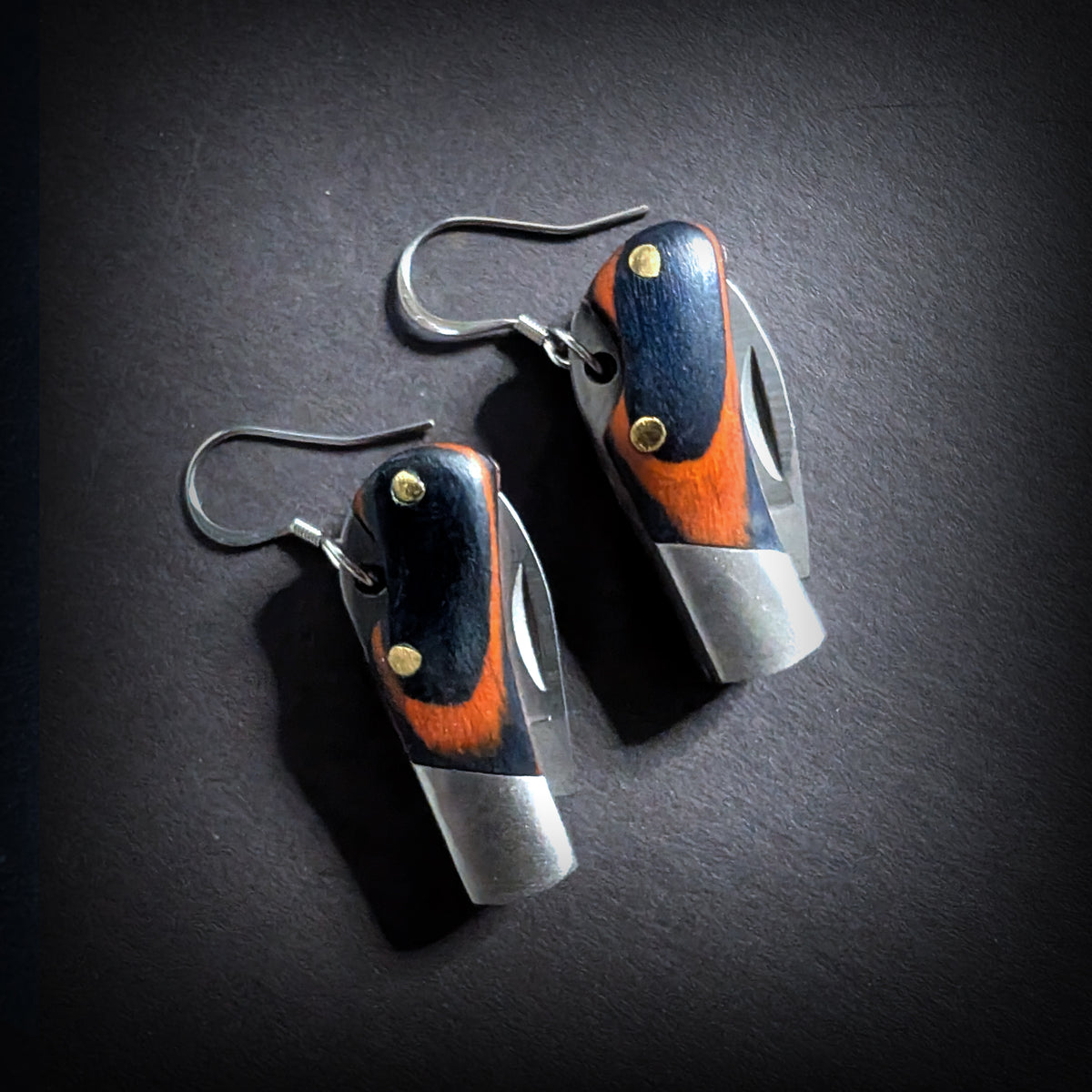 Wooden mini pocket knife earrings by Pocket & Bear Collective, the pair closed on dark grey, showing orange and dark blue wood handles with brass pins and stainless steel bolsters