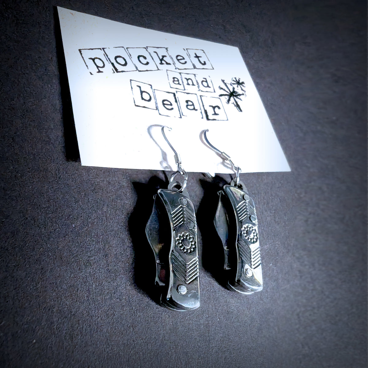 Silver flower mini pocket knife earrings by Pocket & Bear Collective hanging from a stamped Pocket and Bear card on dark grey, both blades closed