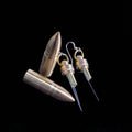 Bullet mini knife earrings by Pocket & Bear Collective with both stainless steel blades fully extended from their brass shells, shown close up on black beside two loose brass bullets