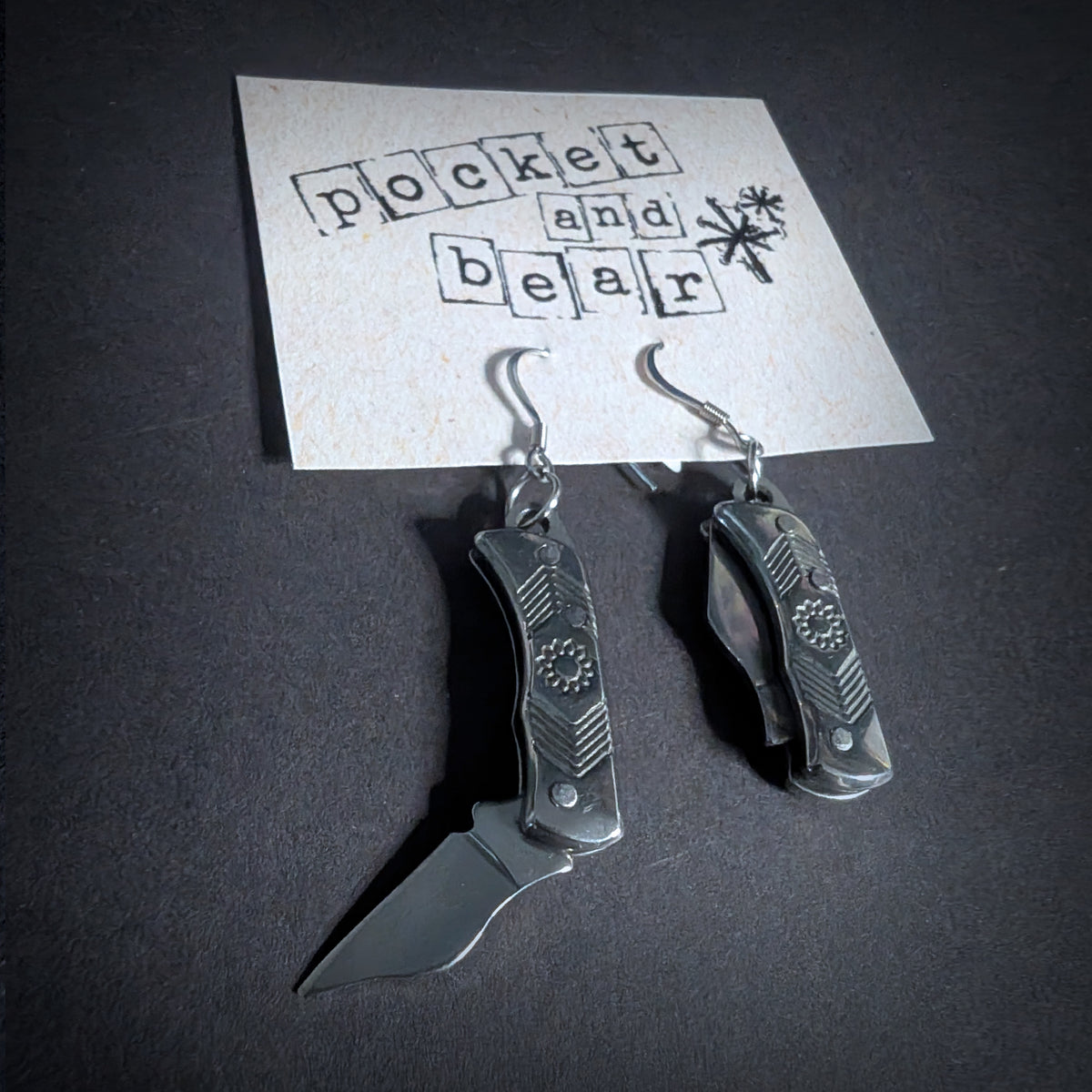 Silver flower mini pocket knife earrings by Pocket & Bear Collective hanging from a stamped Pocket and Bear card on dark grey, one stainless steel blade swung open
