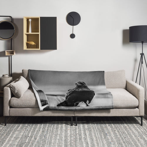 Crow So Chic throw blanket by Iain Camacho, draped over a beige couch in a modern living room with a black and white crow photograph on the front