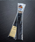 Pack of Arcane Abundance hand dipped incense by Midnight River Apothecary lying at an angle on a dark slate surface, with the label showing at the top of the clear bag and a bundle of black sticks inside