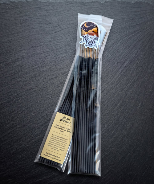 Pack of Arcane Abundance hand dipped incense by Midnight River Apothecary lying at an angle on a dark slate surface, with the label showing at the top of the clear bag and a bundle of black sticks inside