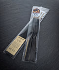 Clear bag of black hand dipped incense sticks with a Midnight River Apothecary Bellus Besom label, lying on a dark slate surface.